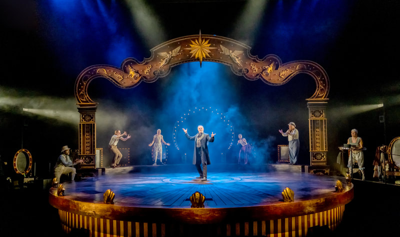 Dramatic smoke- and light-filled circular stage, with performers ranged round the back and an impresario figure at the centre