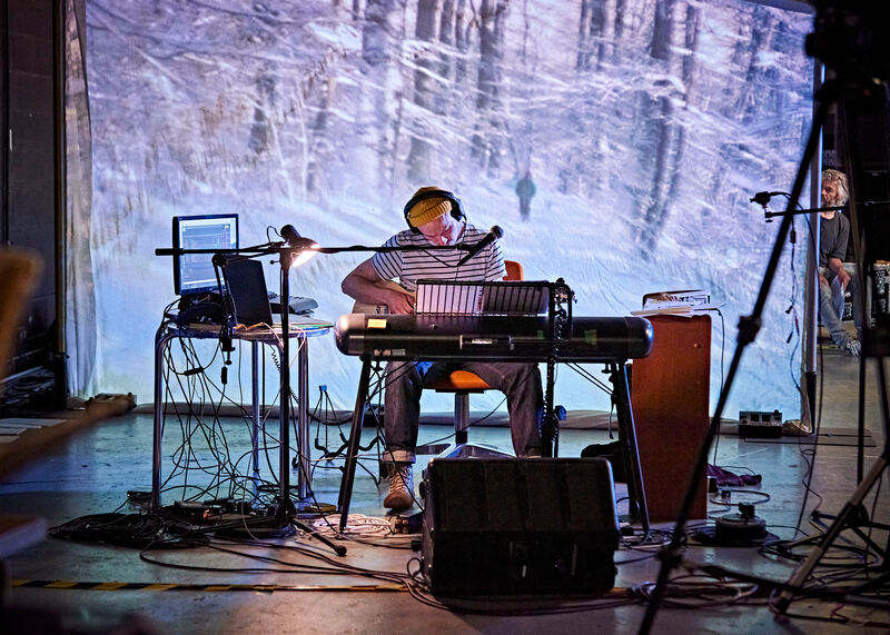 John is sat playing a guitar with computers and keyboards around him, in front of a large projection screen showing snowy woods, and Shôn Dale-Jones in the background