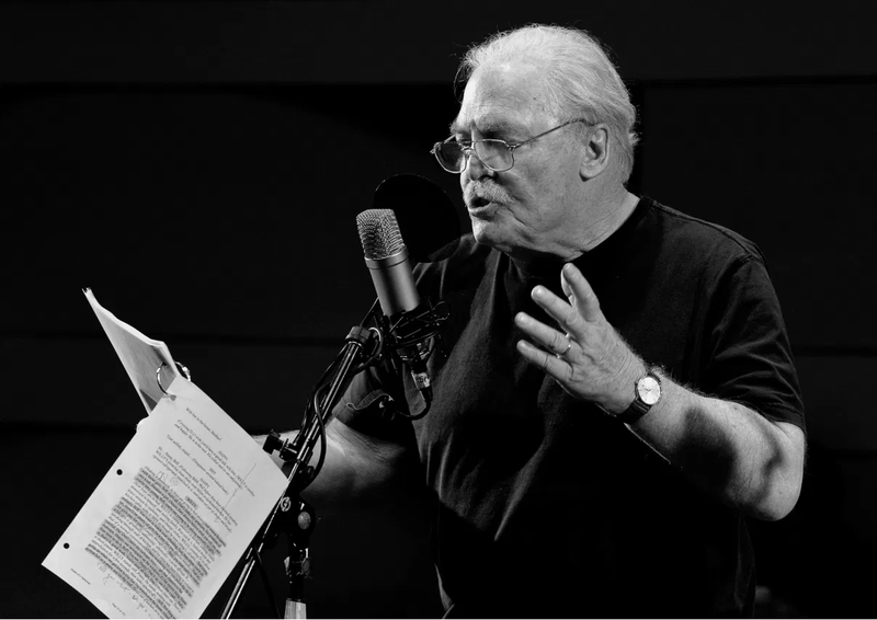 Actor Stacey Keach performing an audio piece into a mic, script in hand
