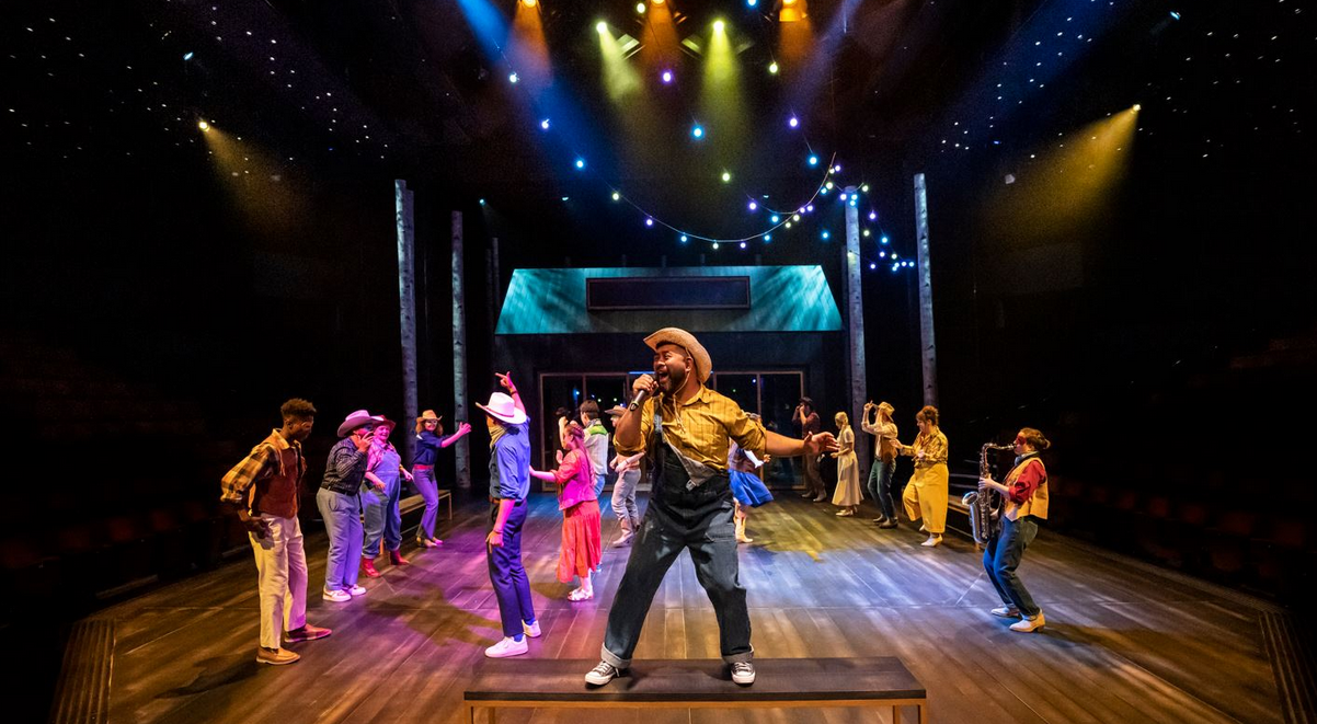 A colourful image of a line dance in full flow, performers all in glamorous cowboy/girl costumes, being led from the foreground by actor Richard Peralta
